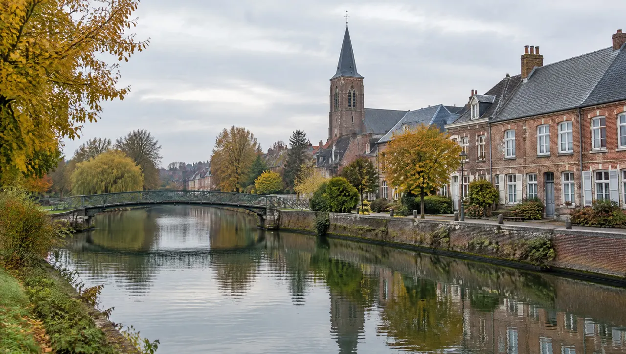 La Lys entre deux quais de briques, une passerelle métallique, des maisons de briques et un clocher sous un ciel gris clair d’automne